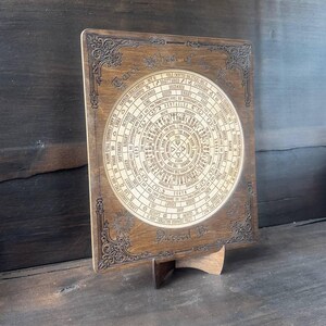 Może przedstawiać: Drewniany wyświetlacz kart tarota z okrągłym wzorem symboli astrologicznych i tekstu. Słowa "Tarot Wheel of the Year" znajdują się na g&oacute;rze, a "Blessed Be" na dole. Wyświetlacz jest podparty drewnianym stojakiem.