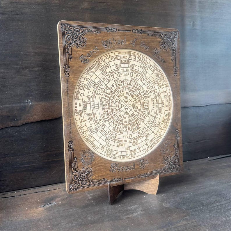Może przedstawiać: Drewniany wyświetlacz kart tarota z okrągłym wzorem symboli astrologicznych i tekstu. Słowa "Tarot Wheel of the Year" znajdują się na g&oacute;rze, a "Blessed Be" na dole. Wyświetlacz jest podparty drewnianym stojakiem.