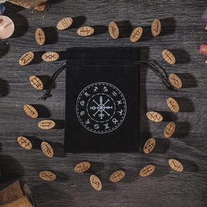 K&ouml;nnte beinhalten: Ein schwarzer Beutel mit Kordelzug, wei&szlig;em kreisf&ouml;rmigem Design und Runensteinen aus Holz. Die Runen sind auf einer dunklen Holzoberfl&auml;che um die Tasche herum verstreut. Die Tasche ist von anderen mystischen Gegenst&auml;nden umgeben.