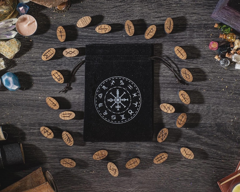 K&ouml;nnte beinhalten: Ein schwarzer Beutel mit Kordelzug, wei&szlig;em kreisf&ouml;rmigem Design und Runensteinen aus Holz. Die Runen sind auf einer dunklen Holzoberfl&auml;che um die Tasche herum verstreut. Die Tasche ist von anderen mystischen Gegenst&auml;nden umgeben.