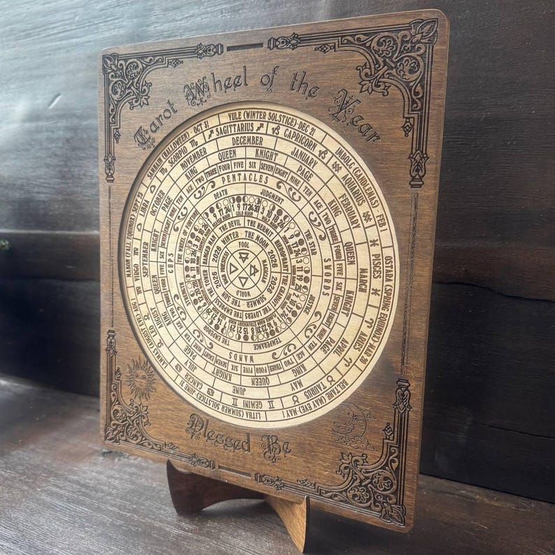 Może przedstawiać: Drewniane Koło Tarota Roku, z okrągłym wzorem z symbolami astrologicznymi, miesiącami i nazwami kart tarota. Ciemnobrązowe drewno ma szczeg&oacute;łowe grawerowane wzory i jest wyświetlane na drewnianym stojaku. Tekst "Blessed Be" znajduje się na dole.