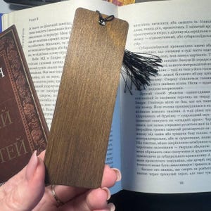 Pu&ograve; includere: Segnalibro in legno marrone scuro con nappa nera. Il segnalibro rettangolare con angoli arrotondati &egrave; posto su un libro aperto.