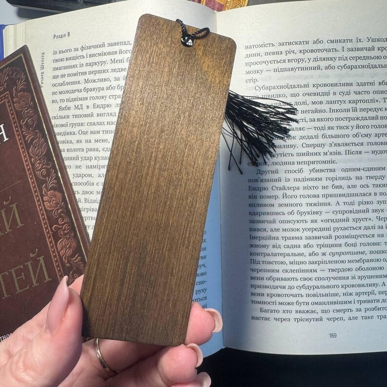 Pu&ograve; includere: Segnalibro in legno marrone scuro con nappa nera. Il segnalibro rettangolare con angoli arrotondati &egrave; posto su un libro aperto.