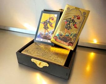May include: A dark wooden box with a gold clasp holds a tarot deck. The deck features gold foil cards with illustrations, including one labeled "THE FOOL." The box and cards are on a reflective surface.
