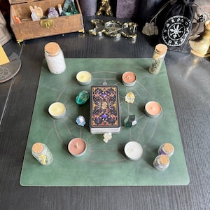May include: A square green mat featuring a circular geometric design, centred with a deck of tarot cards. The mat is surrounded by six tea light candles in various colours, four small glass jars with cork stoppers, and four raw crystals.