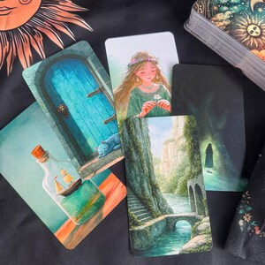 Può includere: Una collezione di carte dei tarocchi con varie illustrazioni, tra cui una nave in una bottiglia di vetro, una porta blu con un gatto, una persona con una corona di fiori, un ponte di pietra sull'acqua e un bosco scuro con una sagoma umana.