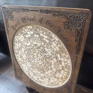 Może przedstawiać: Drewniane Koło Tarota Roku, z okrągłym wzorem z symbolami astrologicznymi, datami i tekstem. Ciemnobrązowe drewno ma dekoracyjne rzeźbienia narożne, a słowa "Tarot Wheel of the Year" są wytrawione.