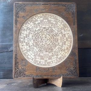 Może przedstawiać: Drewniane Koło Roku Tarota ze skomplikowanymi grawerowanymi detalami. Okrągły wz&oacute;r zawiera symbole astrologiczne, daty i tekst. Rama ma dekoracyjne wzory narożne i słowa "Blessed Be" na dole. Wyświetlane na drewnianym stojaku.