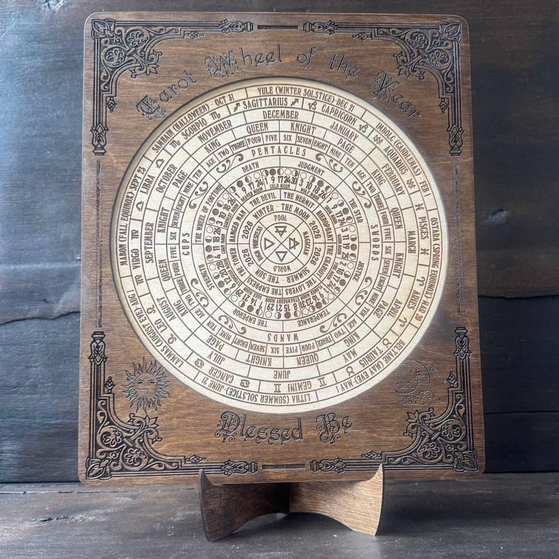 Może przedstawiać: Drewniane Koło Roku Tarota ze skomplikowanymi grawerowanymi detalami. Okrągły wz&oacute;r zawiera symbole astrologiczne, daty i tekst. Rama ma dekoracyjne wzory narożne i słowa "Blessed Be" na dole. Wyświetlane na drewnianym stojaku.