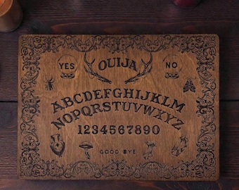 Op de afbeelding: Een rechthoekig houten Ouija-bord met gegraveerde letters, cijfers en symbolen. Het bord bevat het woord "Ouija" en de woorden "Ja" en "Nee". Decoratieve krullen omlijsten de randen van het bord. Het bord is bruin.