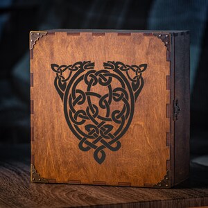 May include: A wooden box with a dark stain and a Celtic knot design. The box has metal corner protectors and a metal clasp. The Celtic knot design is black and features two animal heads at the top.