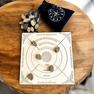 K&ouml;nnte beinhalten: Ein h&ouml;lzernes Divinationsbrett mit eingravierten Texten und Symbolen, darunter "Place of Receiving" und "Ring of Manifestation". Das Brett hat mehrere Runensteine aus Holz und einen schwarzen Samtbeutel mit wei&szlig;em Design. Eine Sammlung von Holzrunen ist ebenfalls vorhanden.