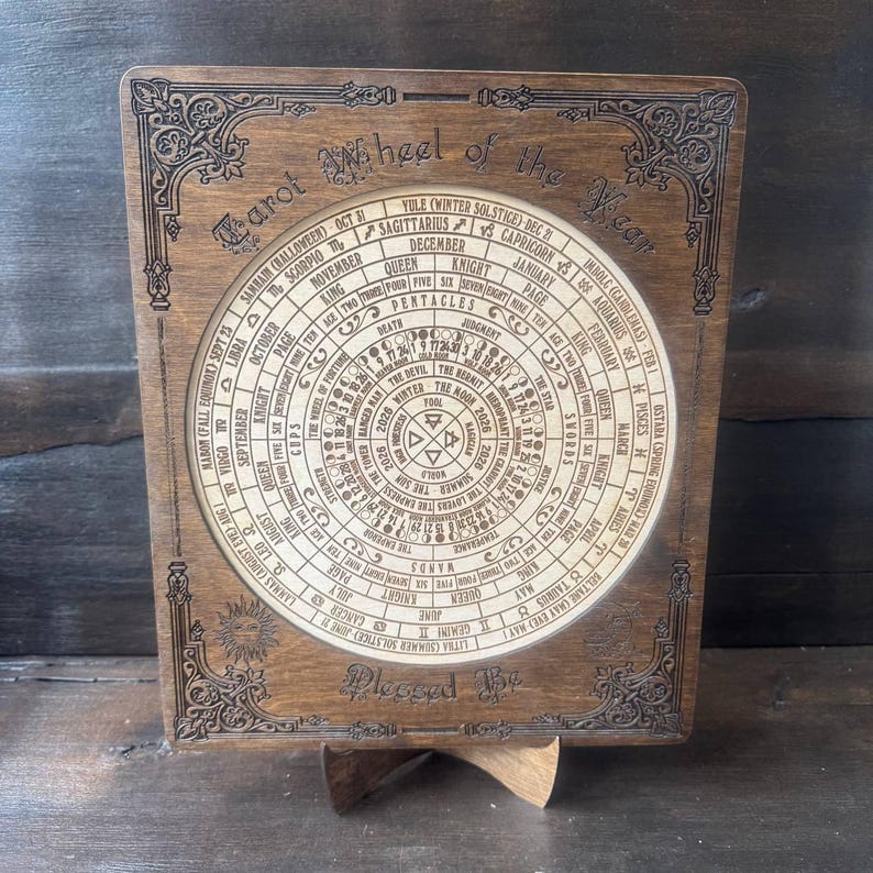 Może przedstawiać: Drewniane koło Tarota Roku z grawerowanymi tekstami i wzorami. Okrągły środek wyświetla symbole astrologiczne i daty, otoczone ozdobną obw&oacute;dką. Tekst "Tarot Wheel of the Year" znajduje się na g&oacute;rze, a "Pleased By" na dole.