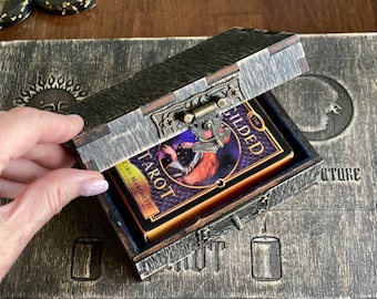 May include: A dark wooden box with a metal latch containing a deck of The Gilded Tarot cards by Ciro Marchetti. The box lid is partially open, revealing the deck inside, which features a purple and gold design with an illustration of a figure.