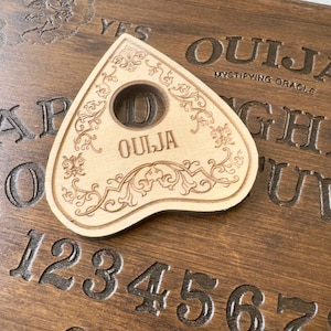 May include: A wooden Ouija planchette with floral designs and the word "OUJIA" rests on a dark brown Ouija board. The board has engraved letters, numbers, and the word "YES". The planchette features a circular opening.