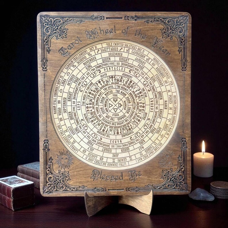 Może przedstawiać: Drewniane Koło Roku Tarota ze skomplikowanymi grawerunkami. Okrągły wz&oacute;r zawiera symbole astrologiczne, nazwy kart i odniesienia sezonowe. Fraza "Blessed Be" jest wpisana na dole. Świeca i inne przedmioty znajdują się w tle.