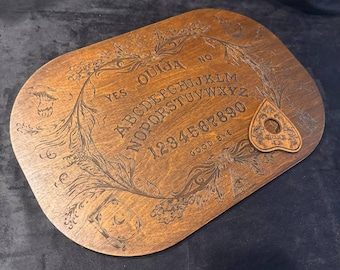 Könnte beinhalten: Ein ovales Ouija-Brett aus Holz mit eingravierten Buchstaben des Alphabets, Zahlen von null bis neun, den Wörtern YES, NO und GOOD BYE sowie dekorativen Abbildungen von Pilzen, Blattwerk und einem Augensymbol, inklusive einer Planchette.