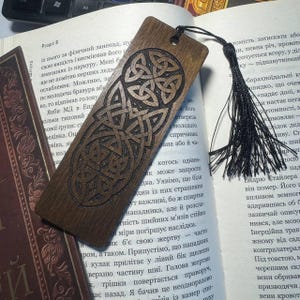 Pu&ograve; includere: Un segnalibro in legno con un disegno a nodo celtico, con finitura marrone scuro. Il segnalibro ha un nappa nera attaccata ed &egrave; appoggiato sulle pagine di un libro. Il segnalibro misura circa 18 cm di lunghezza.