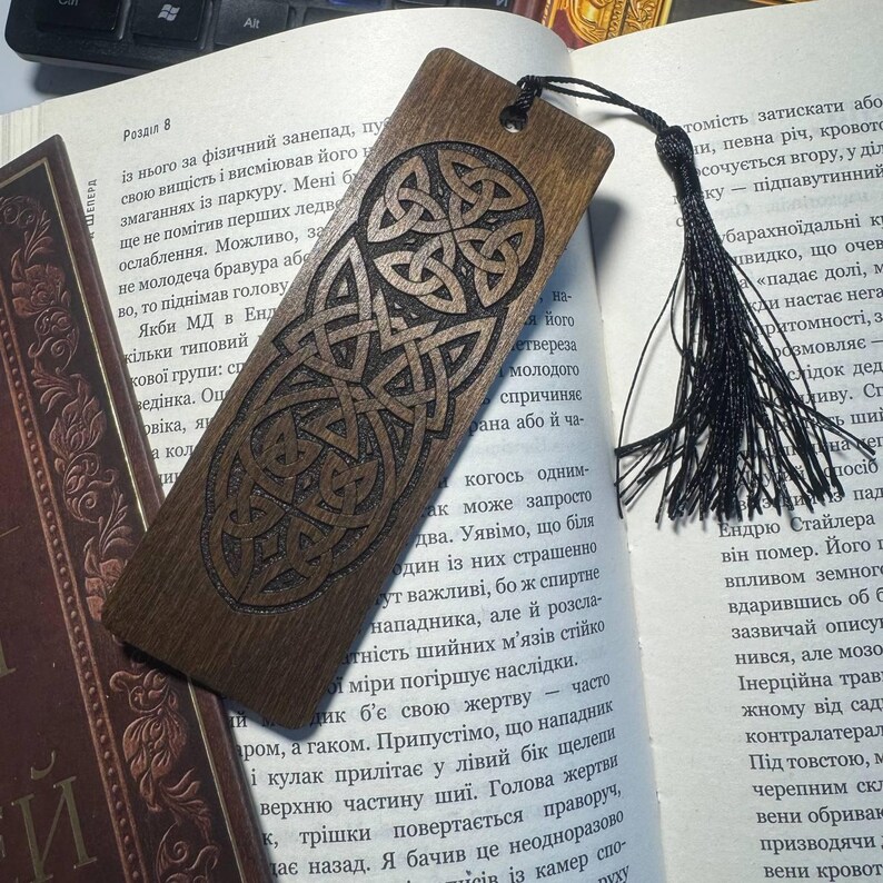 Pu&ograve; includere: Un segnalibro in legno con un disegno a nodo celtico, con finitura marrone scuro. Il segnalibro ha un nappa nera attaccata ed &egrave; appoggiato sulle pagine di un libro. Il segnalibro misura circa 18 cm di lunghezza.