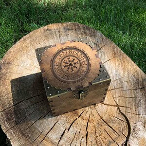 May include: A square wooden box with a dark brown finish, featuring brass-toned metal corner protectors and a front latch. The lid displays a circular design with a central Vegvisir symbol surrounded by a ring of runic characters and Latin text.