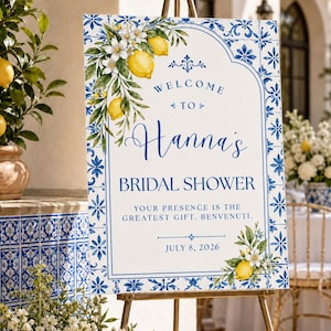 May include: A rectangular welcome sign for a bridal shower featuring a blue and white tile-patterned border. The sign displays the text Welcome to Hanna's Bridal Shower, Your presence is the greatest gift. Benvenuti, and July 8, 2026, with lemon illustrations.