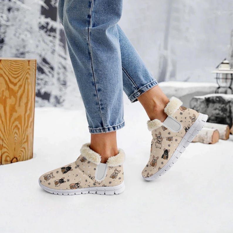 May include: Tan slip-on sneakers with a repeating pattern of French bulldogs in various colors. The shoes have white soles and a fluffy, cream-colored trim around the ankles. The person is wearing blue jeans.