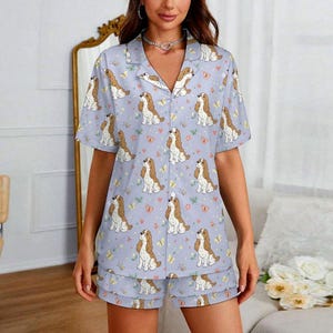 Könnte beinhalten: Hellblaues Pyjama-Set mit einem sich wiederholenden Muster aus braunen und weißen Hunden, Schmetterlingen und Herzen. Das Set beinhaltet ein kurzärmeliges Hemd mit Knöpfen und passende Shorts. Der Pyjama ist aus einem weichen, leichten Stoff gefertigt.