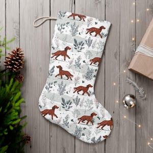 May include: A festive Christmas stocking featuring a repeating pattern of brown dogs, winter foliage, and snow on a white backdrop. It has a cream loop for hanging. The stocking is made of fabric.