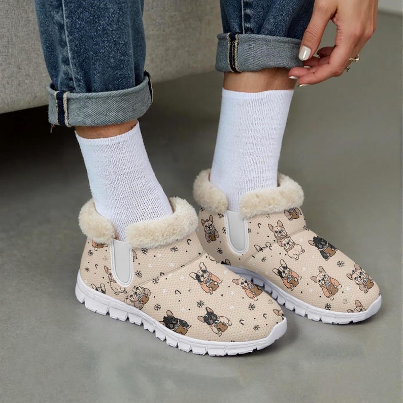 May include: Beige slip-on house shoes with a repeating pattern of French bulldog illustrations in various colors. The shoes have a white sole and a fluffy, cream-colored trim around the opening. The design includes small snowflakes and horseshoes.
