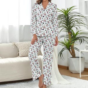 Pode incluir: Conjunto de pijama branco com um padrão repetido de um cão com um casaco vermelho e árvores de Natal verdes. O conjunto inclui uma camisa com botões e calças a condizer. O pijama tem botões pretos e gola.