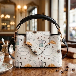 May include: A structured handbag with a cream-colored exterior featuring a repeating pattern of moka pots, croissants, coffee beans, spoons, and the text "coffee time." The bag has a black top handle, gold-tone hardware, and a fold-over flap closure.