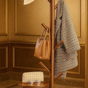 May include: A wooden coat rack with a cream-colored hat, a brown leather handbag, and a black and white patterned coat. A small round table holds a patterned pouch and eyeglasses. The rack is in a room with gold-toned walls.