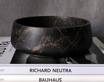 Peut inclure: Un bol rond noir mat orné d'un motif de veines dorées sur toute sa surface extérieure. Le bol est posé sur deux livres empilés aux tranches blanches, l'un portant l'inscription RICHARD NEUTRA et l'autre l'inscription BAUHAUS.