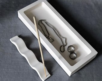 May include: A white rectangular tray containing a silver chain necklace and two silver rings, accompanied by a separate white wavy incense holder with a single incense stick inserted into the end of the holder.