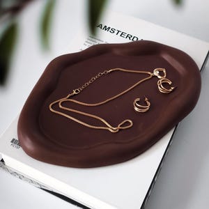 Puede incluir: Una bandeja marrón de forma orgánica con joyas doradas, incluyendo un collar y pendientes de aro. La bandeja descansa sobre un libro blanco con la palabra "AMSTERDAM" visible en la portada. Las joyas se exhiben en la bandeja.