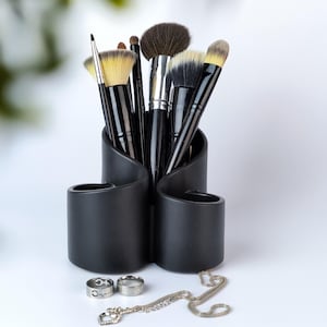 Puede incluir: Un soporte de brochas de maquillaje negro con varios compartimentos, que contiene una variedad de brochas con mangos negros y cerdas naturales. El soporte está sobre una superficie blanca.