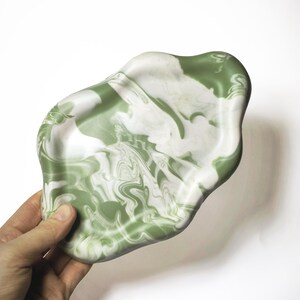 Peut inclure: Un plateau en céramique marbré vert et blanc avec un bord irrégulier et ondulé. Le plateau est fait d'un matériau lisse et brillant.