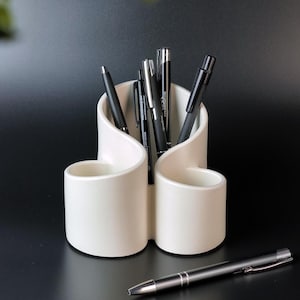 Puede incluir: Organizador de escritorio de cerámica blanca con un diseño de onda único. El organizador tiene tres compartimentos para bolígrafos, lápices u otros artículos de oficina. Hay bolígrafos negros en el organizador.