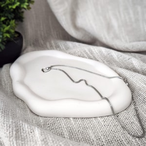 Może przedstawiać: Srebrny łańcuszek z małym serduszkowym wisiorkiem leży na białej ceramicznej tacy z falistym brzegiem. Taca znajduje się na jasnoszarym materiale.