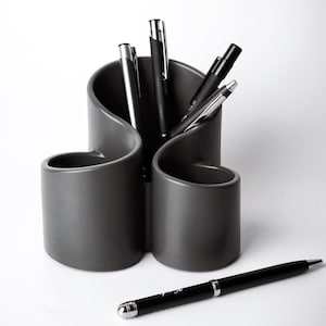 Könnte beinhalten: Ein dunkelgrauer Stifthalter mit einem einzigartigen, geschwungenen Design, der mehrere Stifte enthält. Die Stifte sind schwarz und silberfarben, wobei ein Stift horizontal vor dem Halter liegt. Der Stifthalter besteht aus einem glatten, matten Material.