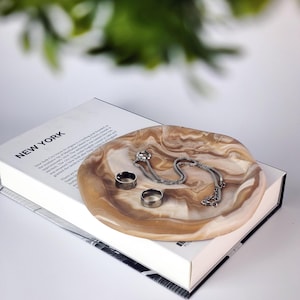 Op de afbeelding: Een bruine en witte marmeren keramische schaal met een zilveren kettingketting en twee zilveren ringen erop. De schaal ligt op een wit boek met het woord "NEW YORK" op de omslag.