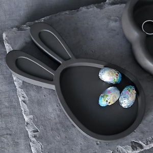 Könnte beinhalten: Ein dunkelgraues, hasenförmiges Tablett mit drei folienverpackten Schokoladeneiern. Das Tablett steht auf einer strukturierten, grauen Oberfläche. Das Design ist minimalistisch und thematisch auf Ostern ausgerichtet.