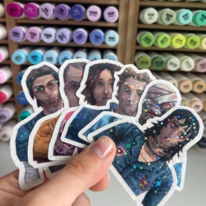Könnte beinhalten: Eine Hand hält eine Sammlung holografischer Aufkleber mit Porträts von Charakteren. Die Aufkleber haben ein glänzendes Finish und sind in verschiedenen Farben gehalten. Der Hintergrund umfasst ein Regal voller bunter Marker.