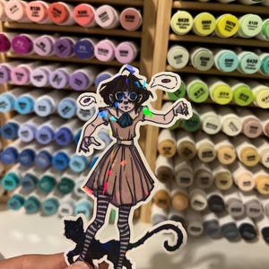 May include: A holographic sticker featuring a cartoon illustration of a person in a dress with striped stockings and a black cat. The sticker has a unique, iridescent finish. The background shows a collection of colorful art markers.