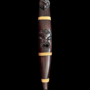 Può includere: Un manufatto in legno intagliato marrone scuro con intricati disegni facciali e accenti turchesi. Il pezzo allungato presenta due volti scolpiti, legati con fasce intrecciate. Il fondo ha una punta con un motivo intagliato. Lo sfondo è nero.