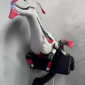 May include: A white creature figurine with a long neck and head, featuring pink accents on the snout and crest, and black and red markings along the body. The creature is mounted on a black, curved base with a dangling red tail-like extension.