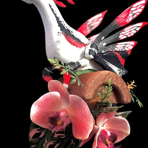 May include: A white creature with black and red markings, featuring wing-like appendages with red and white patterns, perched on a brown cylindrical base. The base is decorated with two large pink orchid flowers and green foliage against a black background.