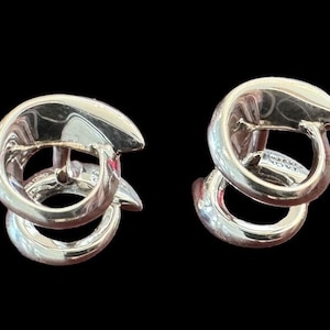 May include: A pair of silver earrings with a double-loop design. The earrings have a polished, reflective surface, with a curved top and a rounded bottom loop. The background is black, which accentuates the silver's shine.