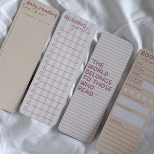 Könnte beinhalten: Vier rechteckige Lesezeichen aus Papier in Creme und Beige mit aufgedrucktem Text. Eines enthält einen täglichen Lesetracker, ein weiteres listet Leseziele auf, eines zeigt das Zitat THE WORLD BELONGS TO THOSE WHO READ und das letzte ist ein Buchbewertungsformular.