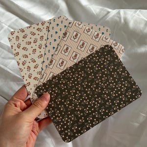 Könnte beinhalten: Ein Set aus vier rechteckigen Karten mit abgerundeten Ecken, die jeweils ein unterschiedliches Muster aufweisen. Die Designs umfassen kleine rostfarbene Blumenmotive, ein blaues geometrisches Gitter, beige Streifen mit Tulpen und einen dunkelbraunen Blumendruck.